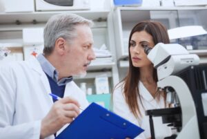 Male scientist in a lab coat coaching female post-doc at a microscope on leadership soft skills